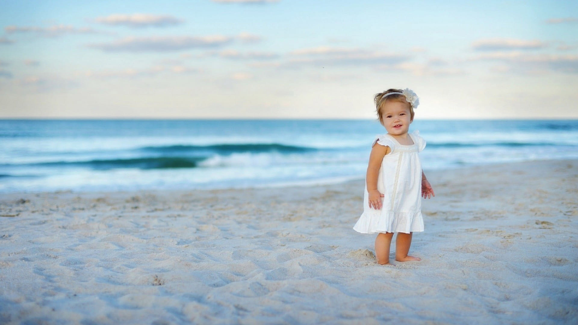 Cute Baby Child Is Standing On Beach Sand Wearing White Dress Under Blue Sky 2K Cute