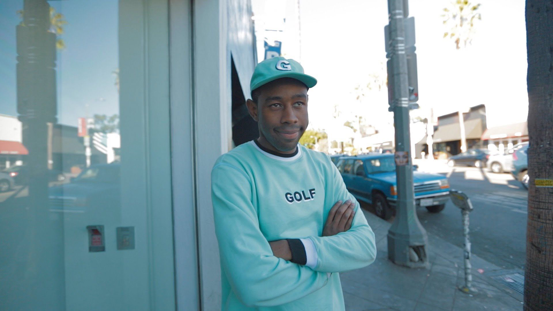 Tyler The Creator Is Smiling And Looking Straight Standing Near Glass Door Wearing Aquamarine Tshirt And Cap 2K Music