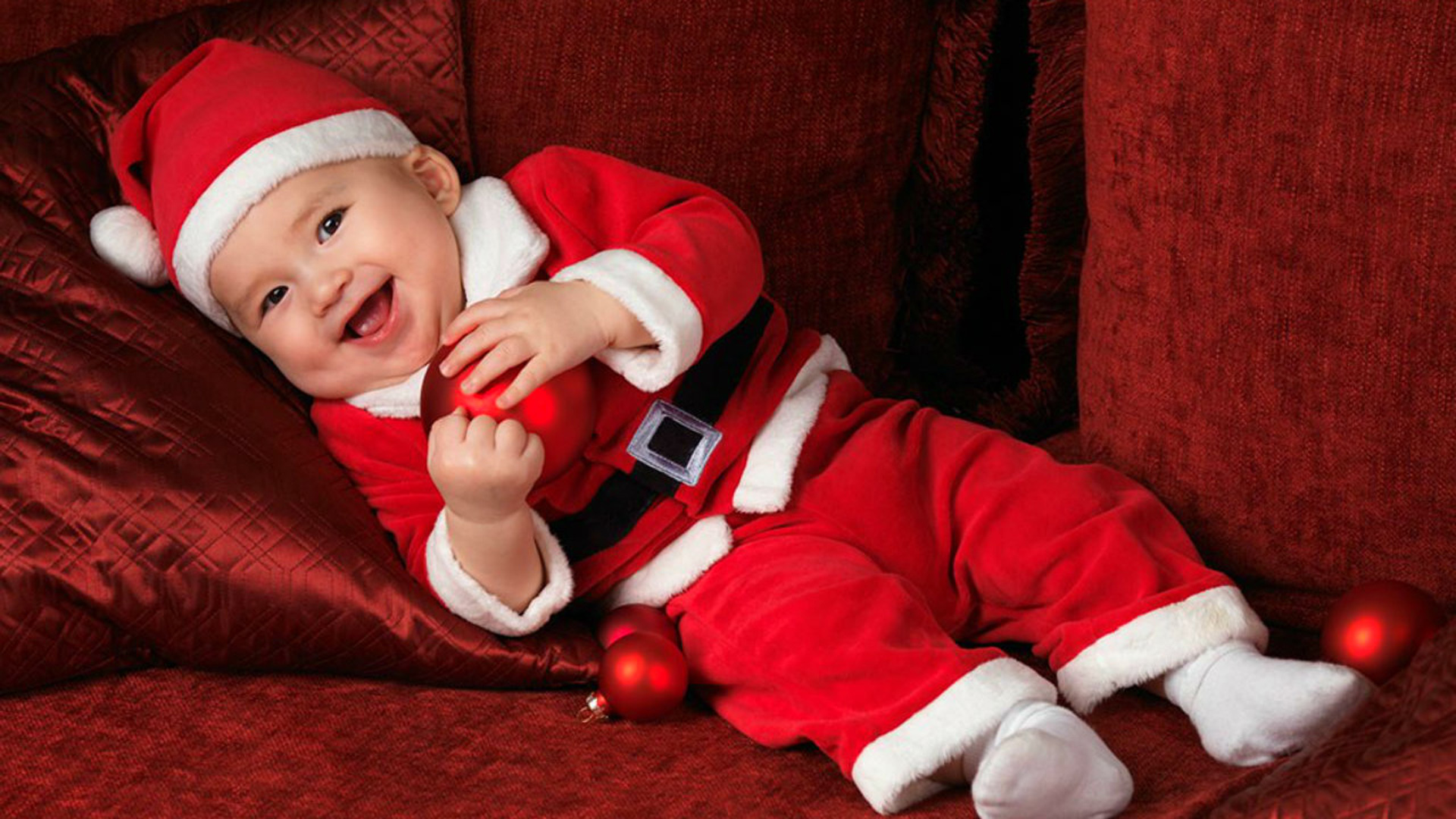 Beautiful Cute Smiling Baby Is Wearing Santa Claus Red Dress Cap Lying Down On Dark Maroon Cloth 2K Cute