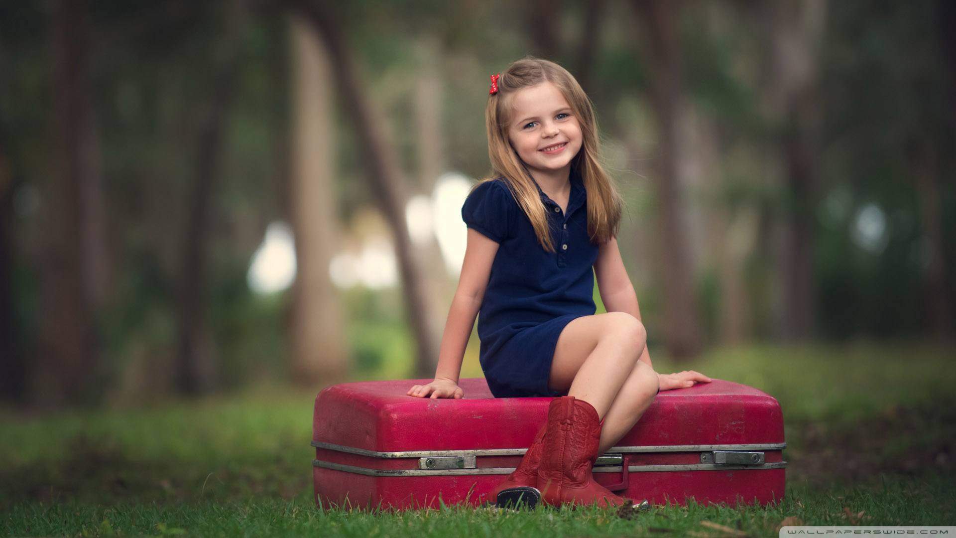 Smiley Cute Little Girl Is Wearing Blue Dress Sitting On Red Suitcase In Blur Trees Wallpaper 2K Cute